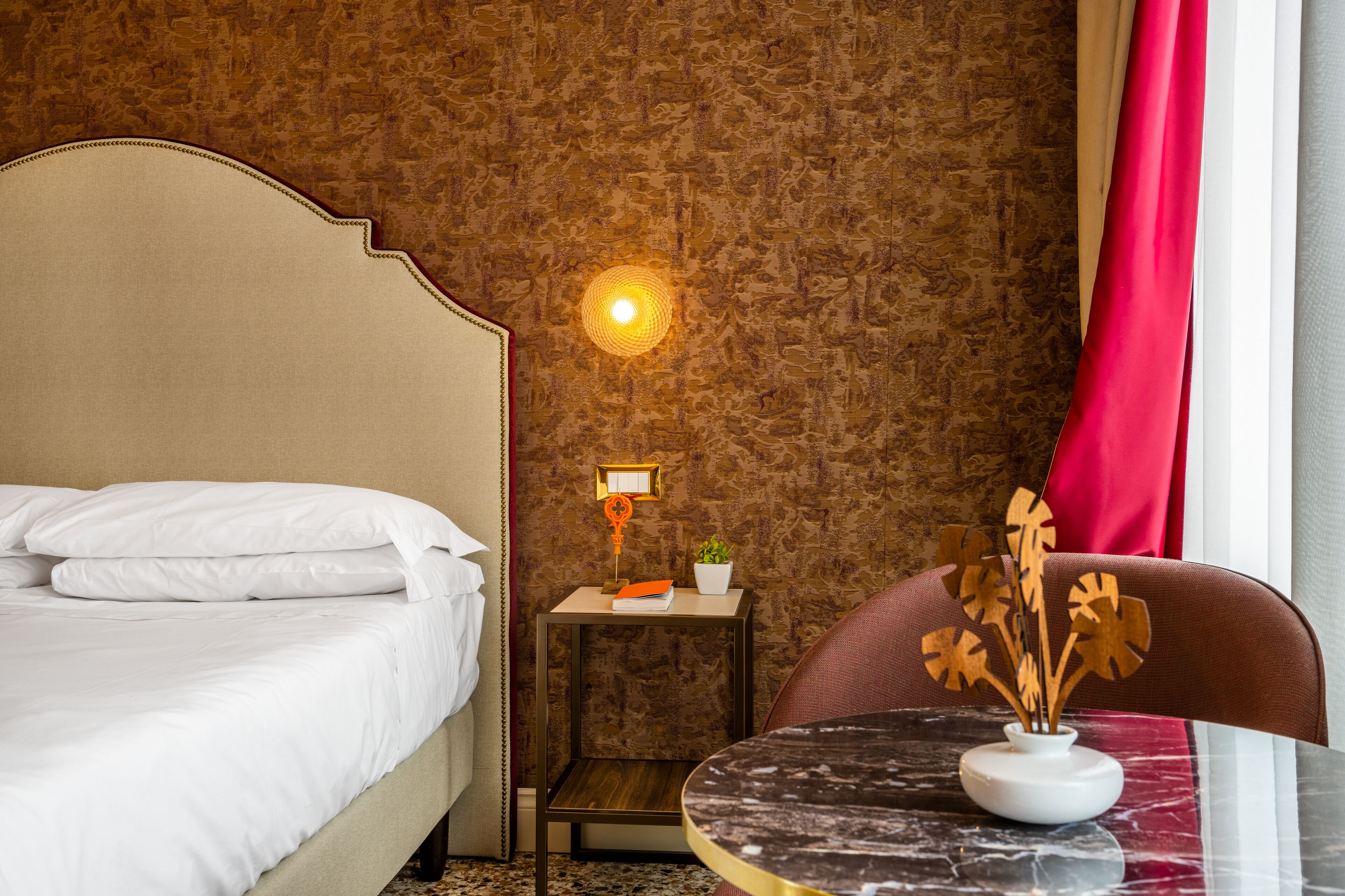 A hotel room with a bed, a nightstand with a lamp, a small table with a vase, and a chair.