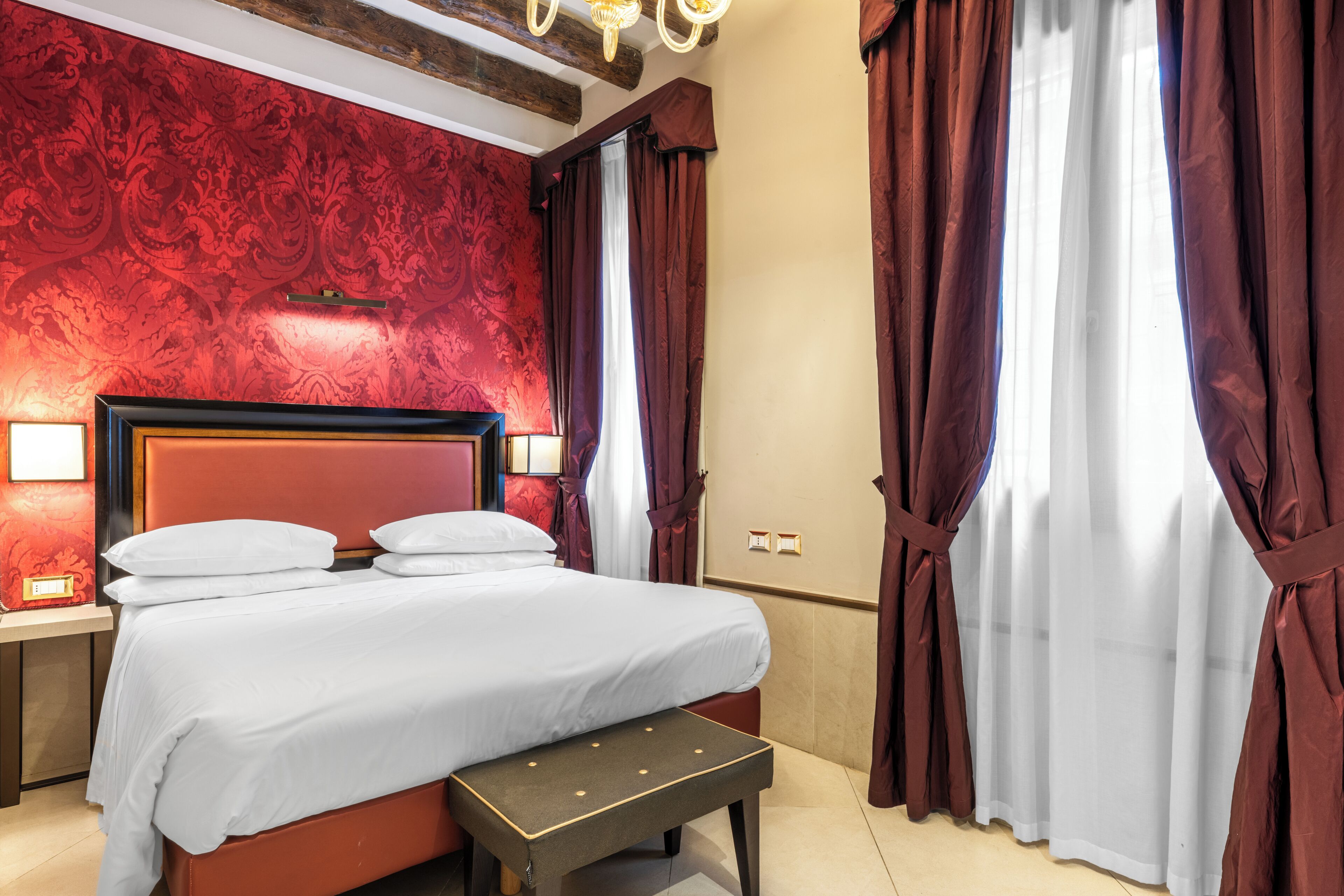A hotel room with a large bed, a bench, a nightstand, and a wall with a red patterned wallpaper.