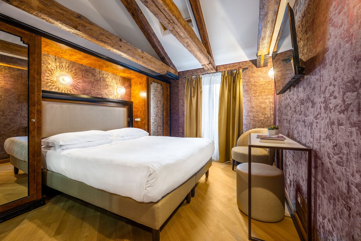 A neatly made bed in a room with exposed wooden beams, a small round table, and a mirror on the wall.