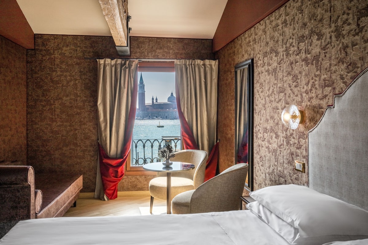 A hotel room with a bed, a small table, two chairs, and a view of a historic building through the window.