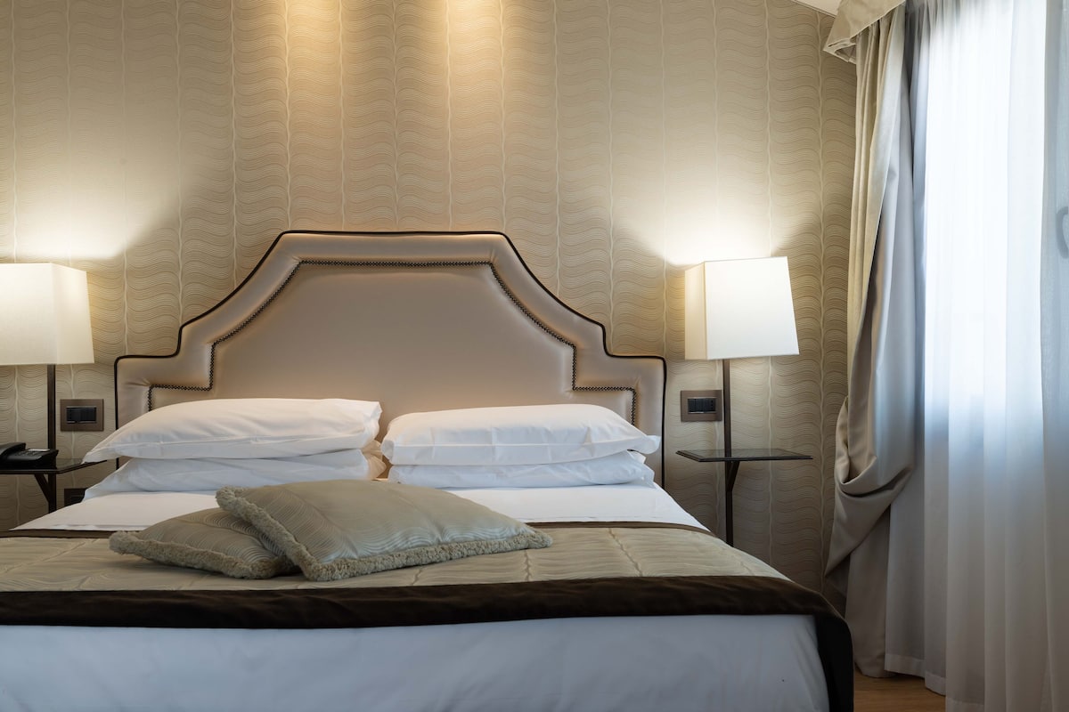 A neatly made bed with a headboard, two bedside lamps, and a window with curtains.