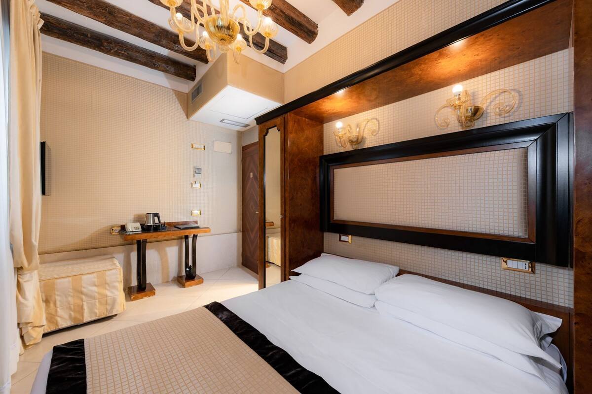 A bedroom with a large bed, a bedside table with a lamp, a wooden headboard, and a mirror.
