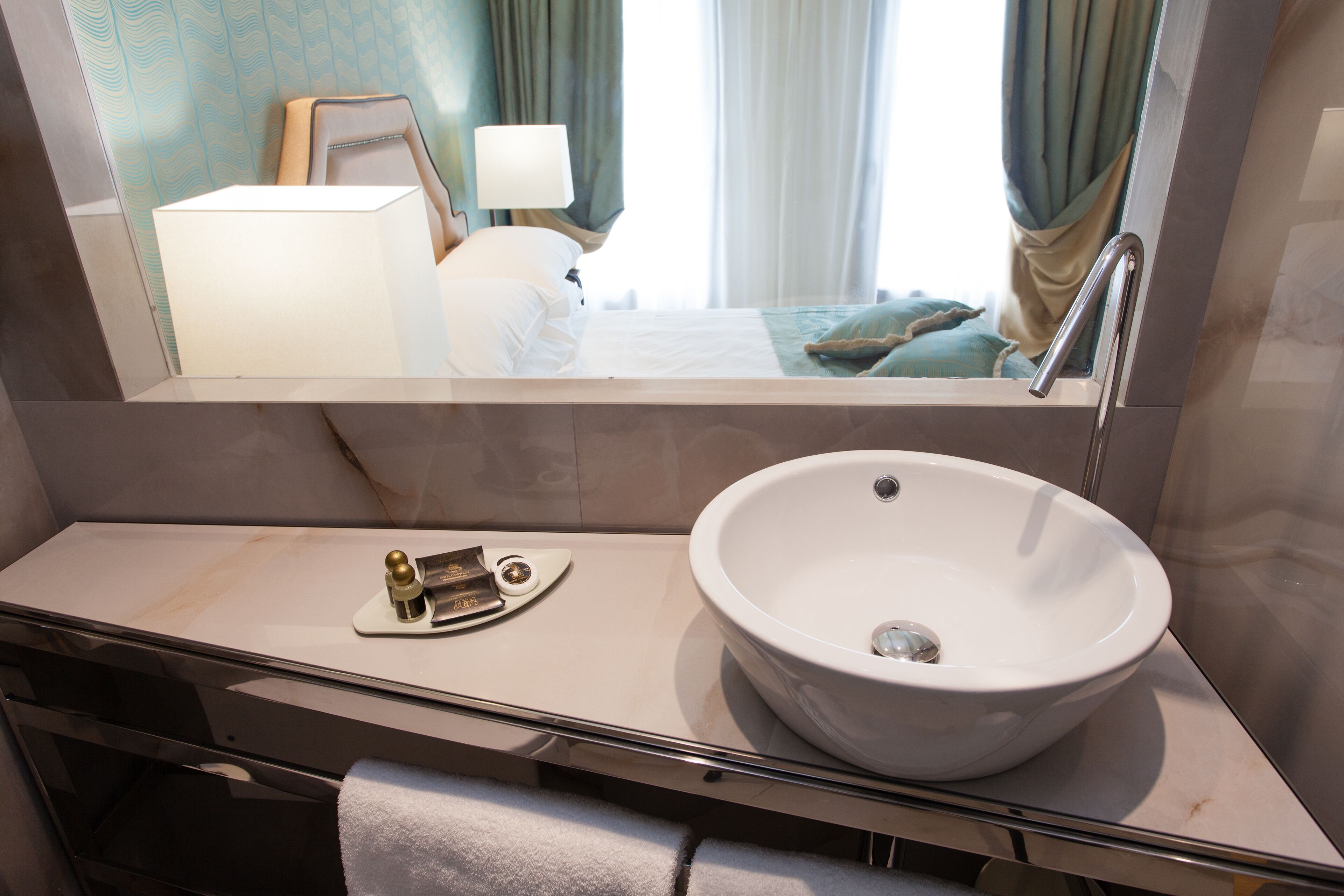 A modern bathroom with a large white sink, a mirror, and a bedside table with a lamp.