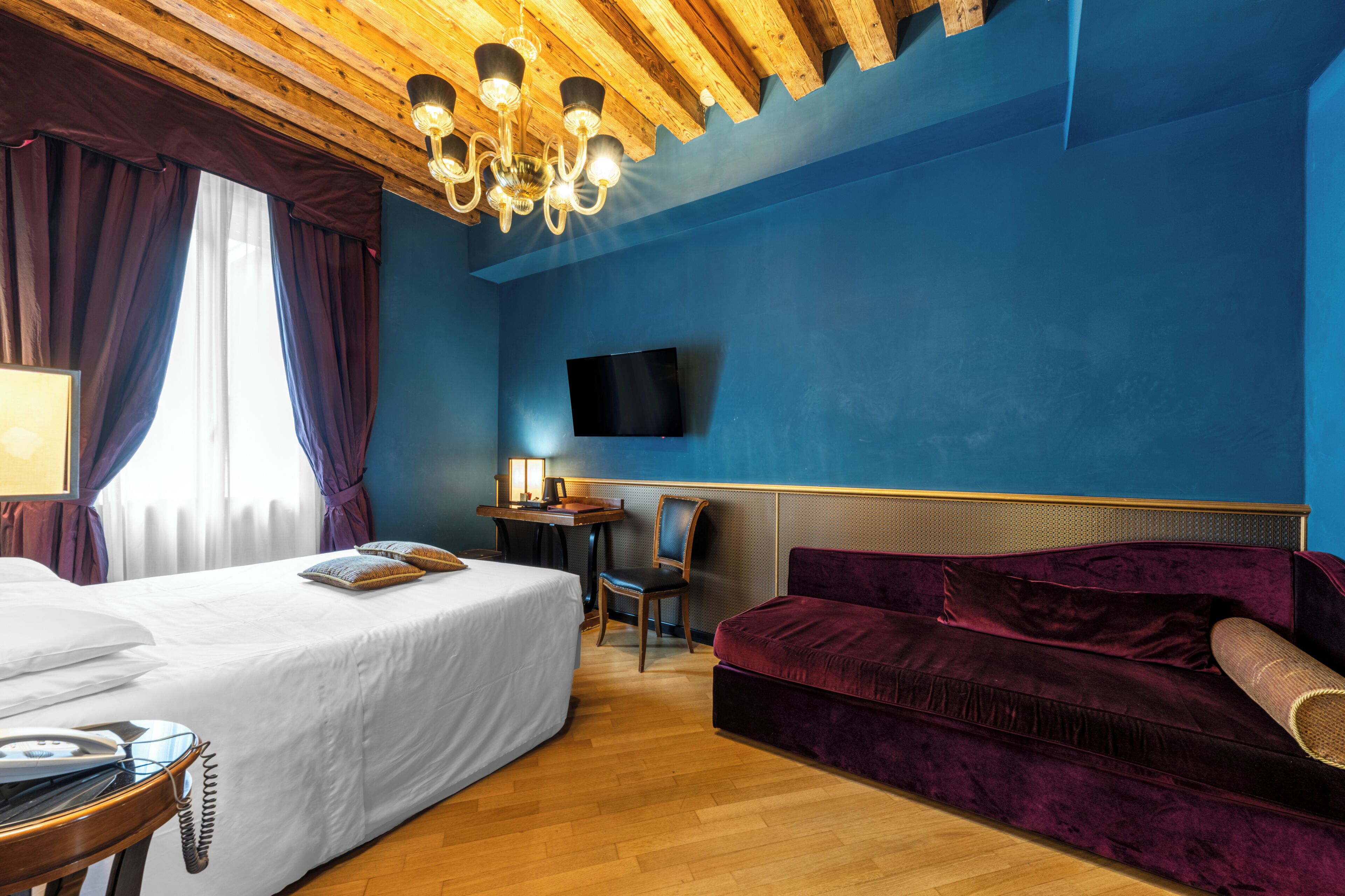 A hotel room with a bed, a sofa, a desk with a chair, a television mounted on the wall, and a wooden ceiling.