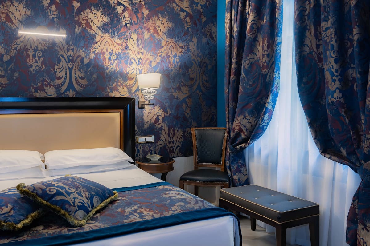 A hotel room with a large bed, a chair, a small table, and a nightstand with a lamp.
