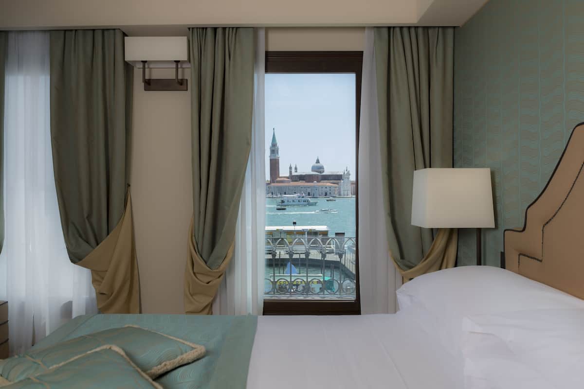 A hotel room with a bed, a nightstand, a lamp, and a view of a cityscape through a window.