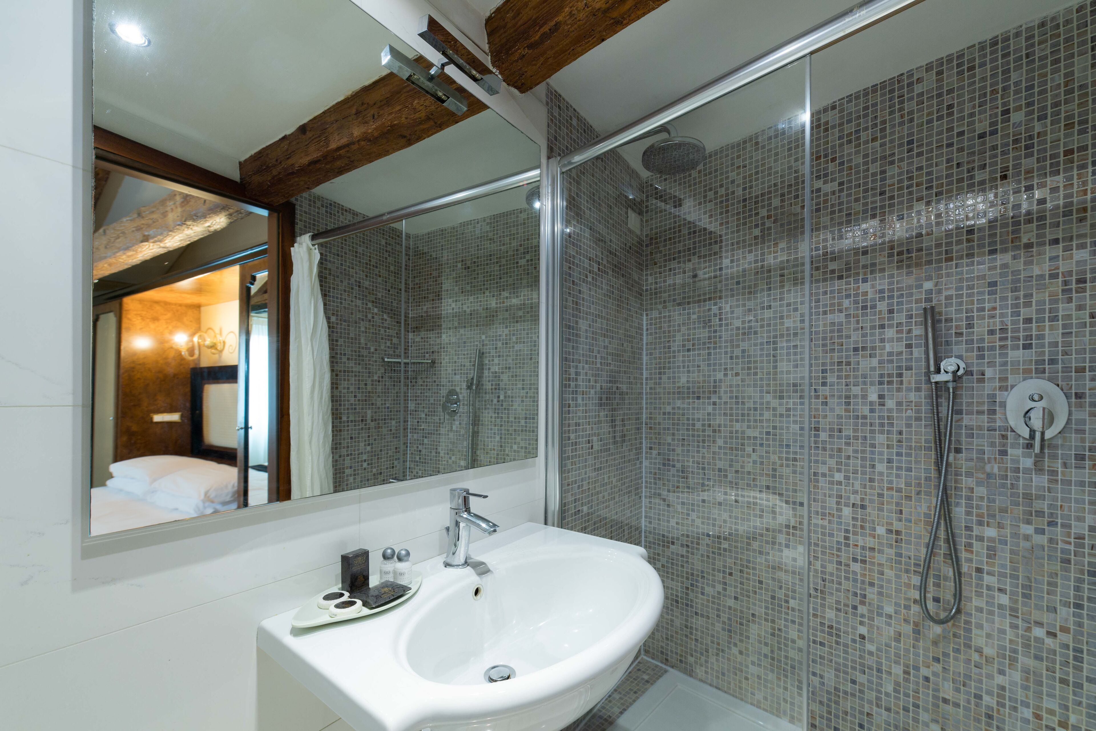 Un baño moderno con un espejo grande, un lavamanos blanco y una zona de ducha con mosaicos.