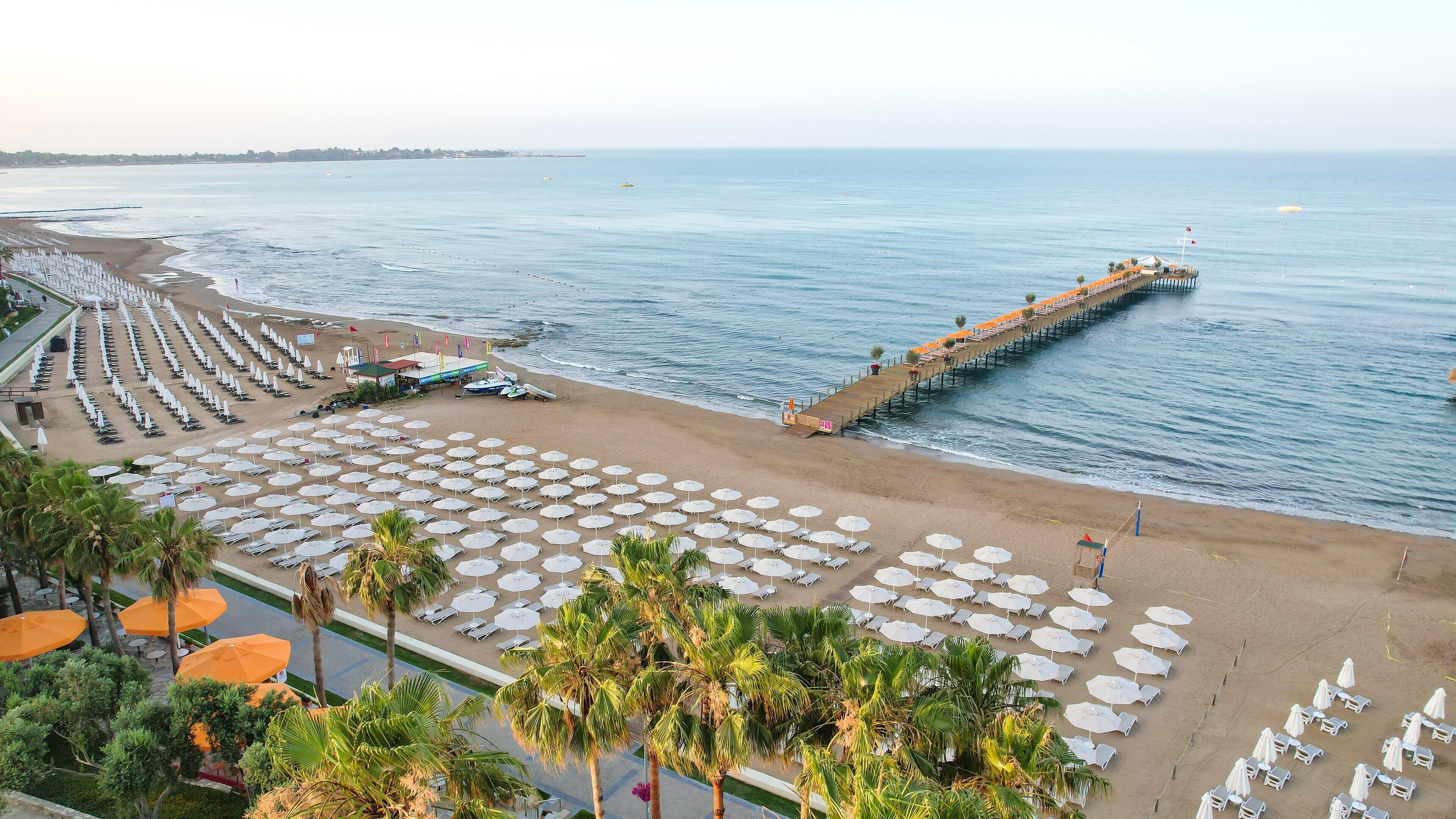 A beach resort with rows of sun loungers and umbrellas, a pier extending into the sea, and a view of the coastline.