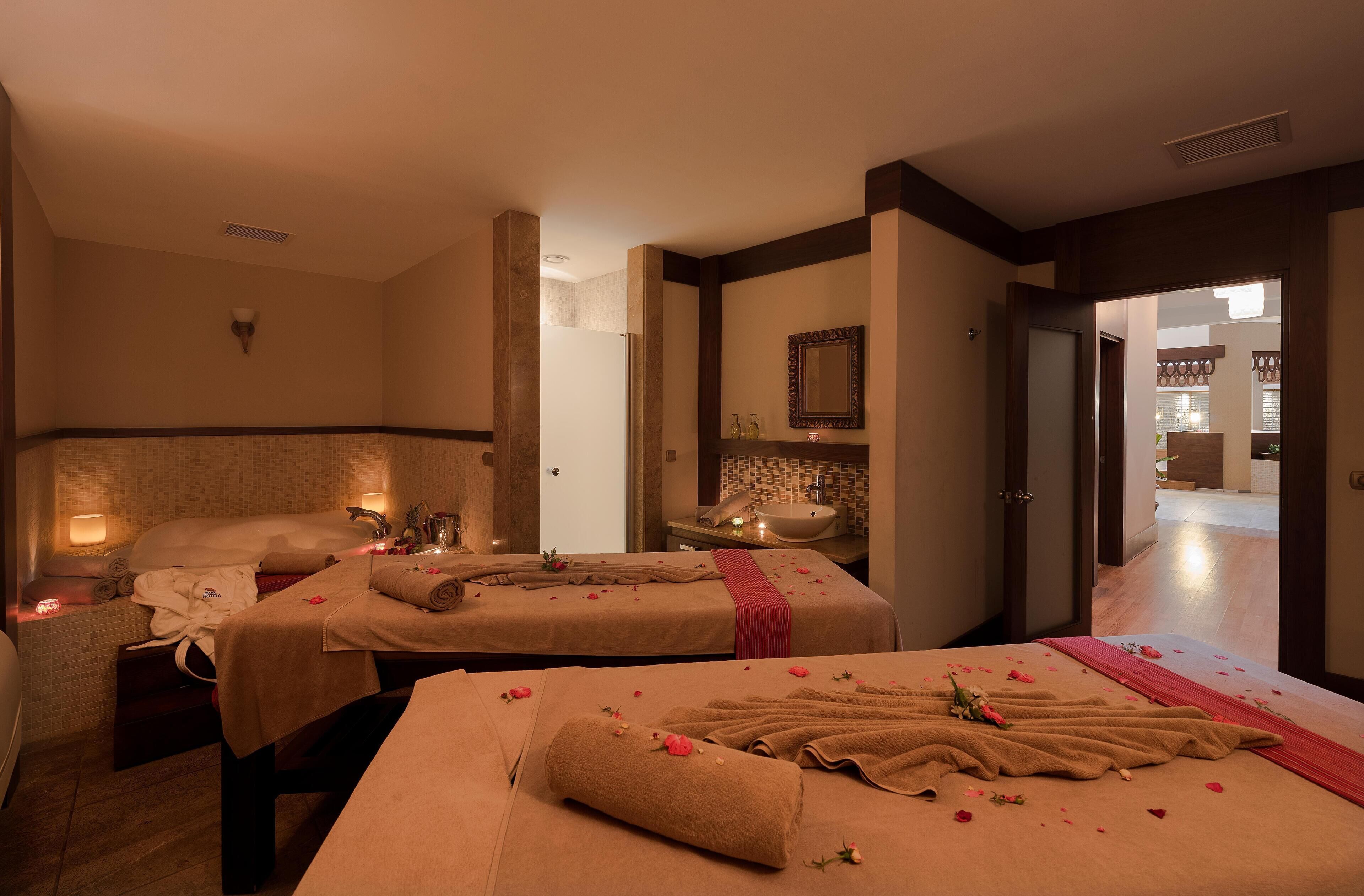 A spa room with two massage tables, each with towels and flowers. There is a sink and a mirror on the wall.