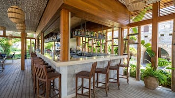 Een houten bar met barkrukken, planken met flessen en uitzicht op groen buiten.