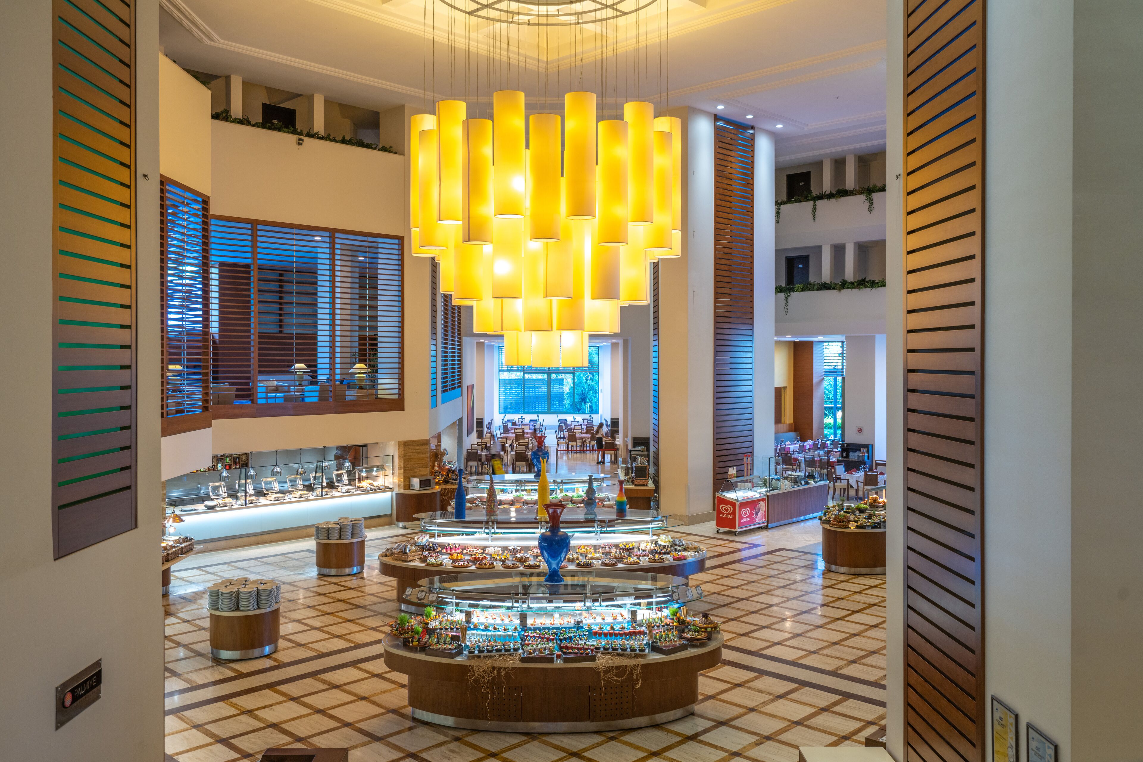 A spacious lobby with a central chandelier, a food display, and a seating area with tables and chairs.