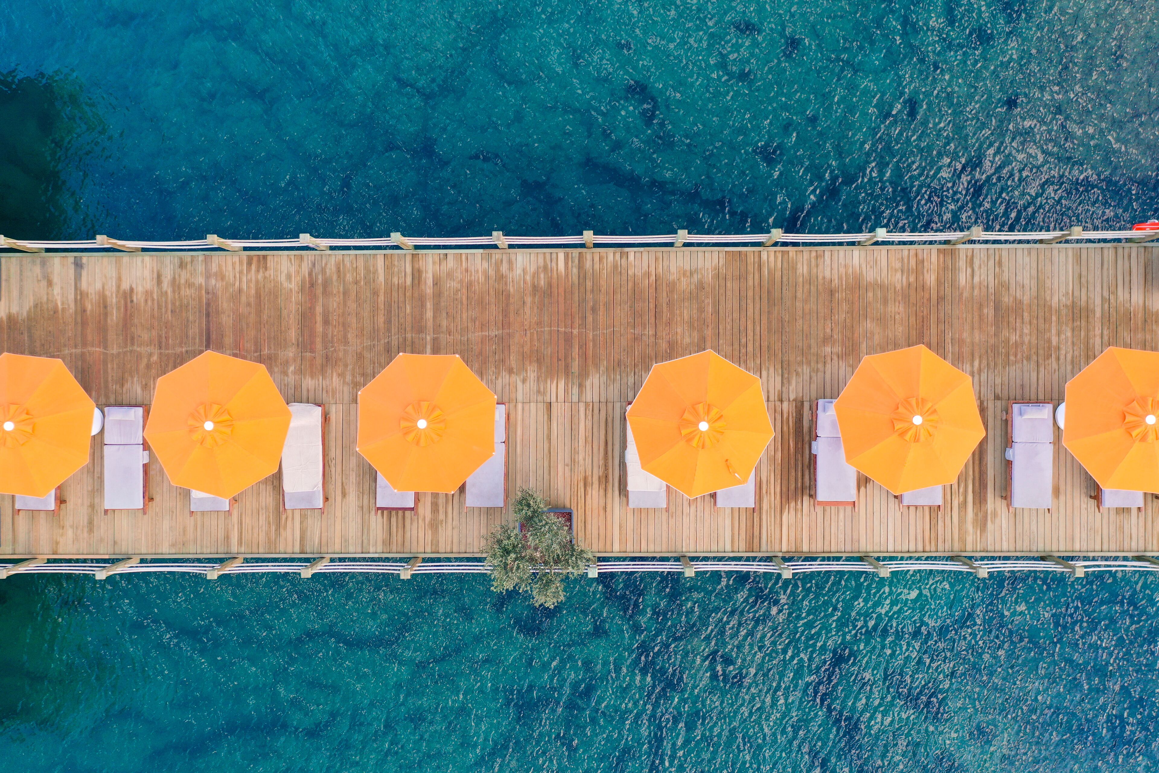 Vue aérienne d’une terrasse en bois avec six chaises longues et six parasols orange, donnant sur une mer d’un bleu limpide.