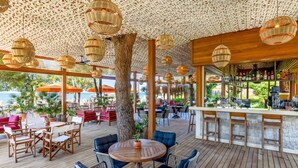 An outdoor seating area with wooden tables and chairs, a large tree, and a bar with a view of the ocean. - Arum Barut Collection - Ultra All Inclusive (Manavgat)