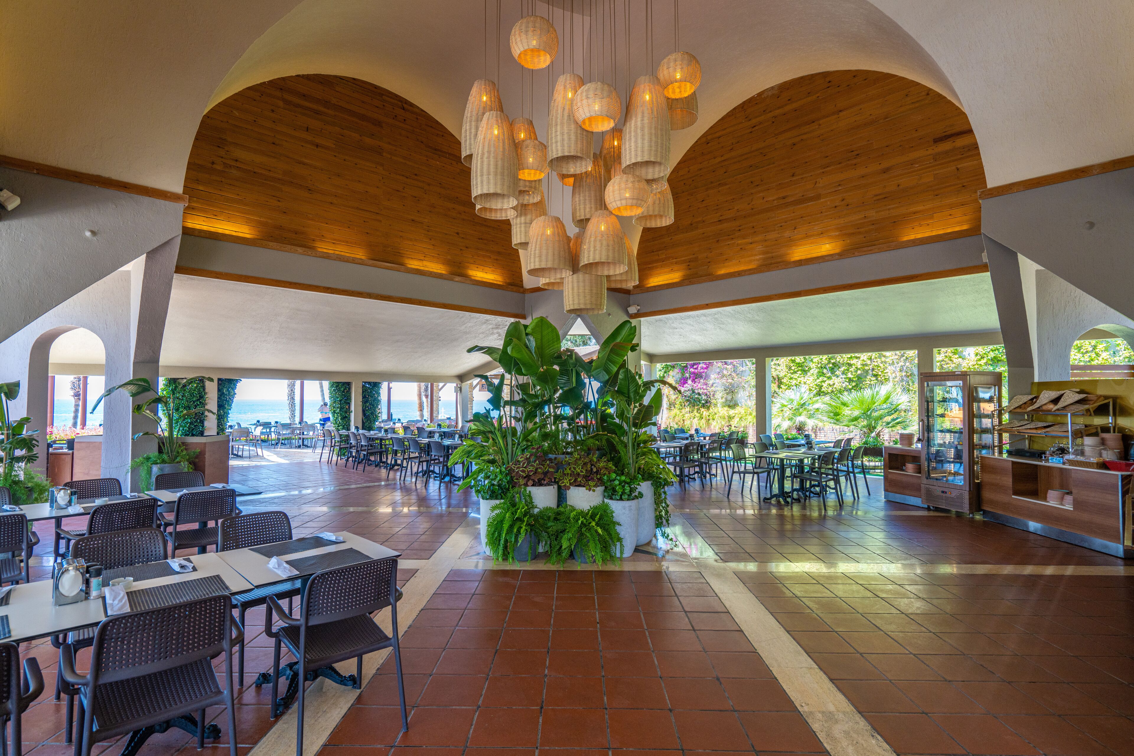 A spacious indoor dining area with high wooden ceilings, multiple tables and chairs, and potted plants.