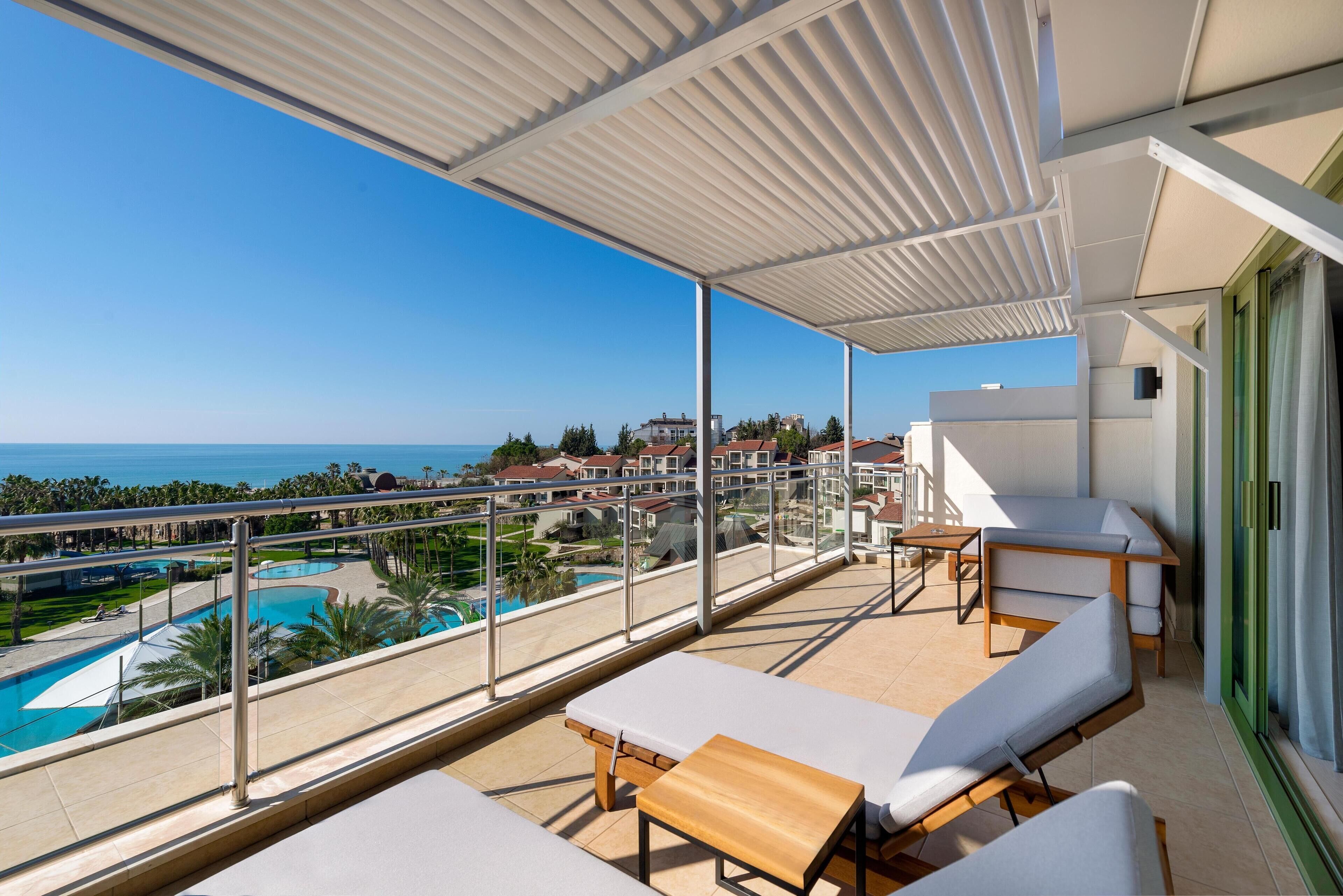 A balcony with white cushioned chairs and a wooden table, overlooking a swimming pool and a clear blue sky.