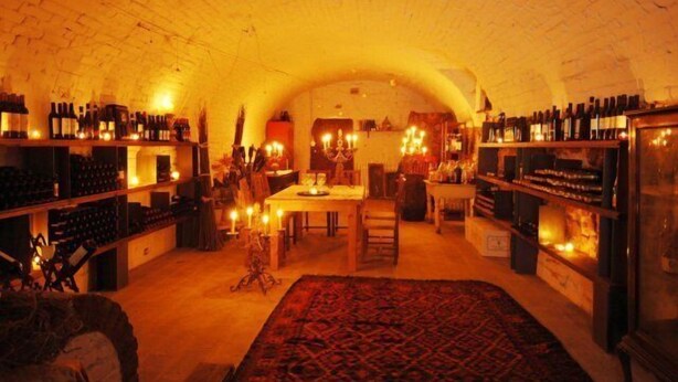 An underground wine cellar with shelves of wine bottles, a dining area with a table and chairs, and a patterned rug on the floor. - Relais del Castello di Oviglio (Oviglio)