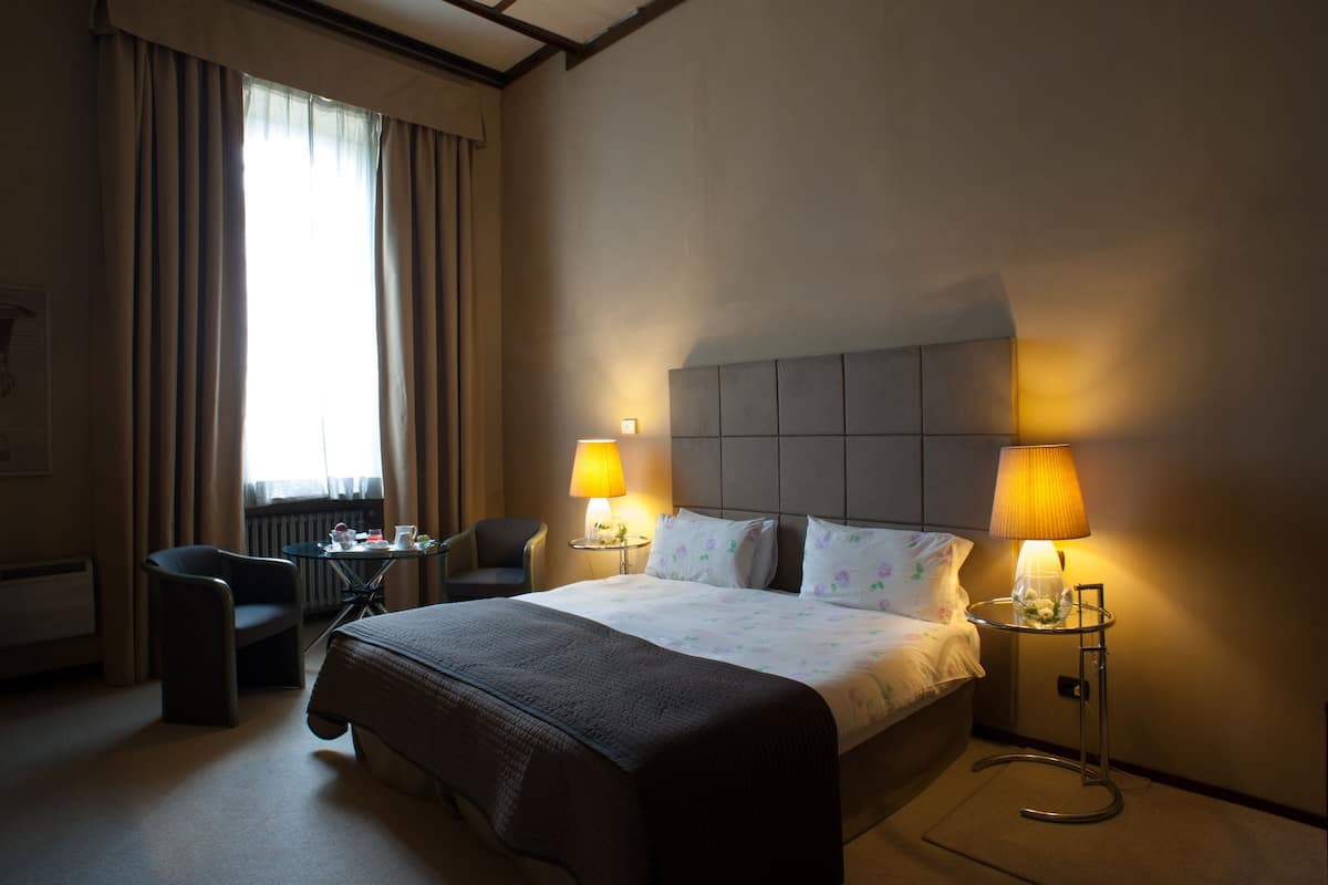 A hotel room with a large bed, two bedside lamps, a small table, a chair, and a window with curtains.
