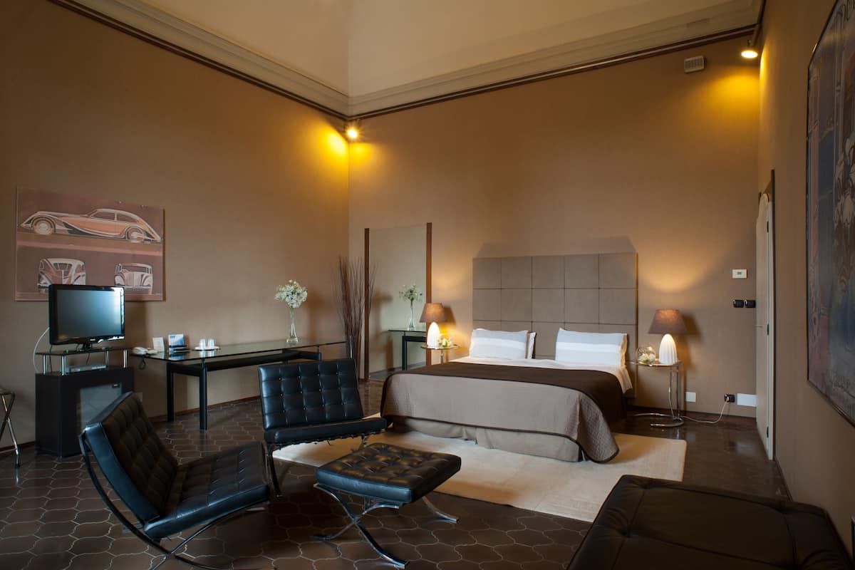 A hotel room with a bed, two chairs, a desk with a computer, a television, and a painting on the wall.