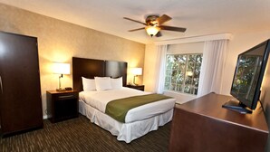 A hotel room with a large bed, two bedside tables with lamps, a ceiling fan, a TV on a wooden stand, and a window with curtains.