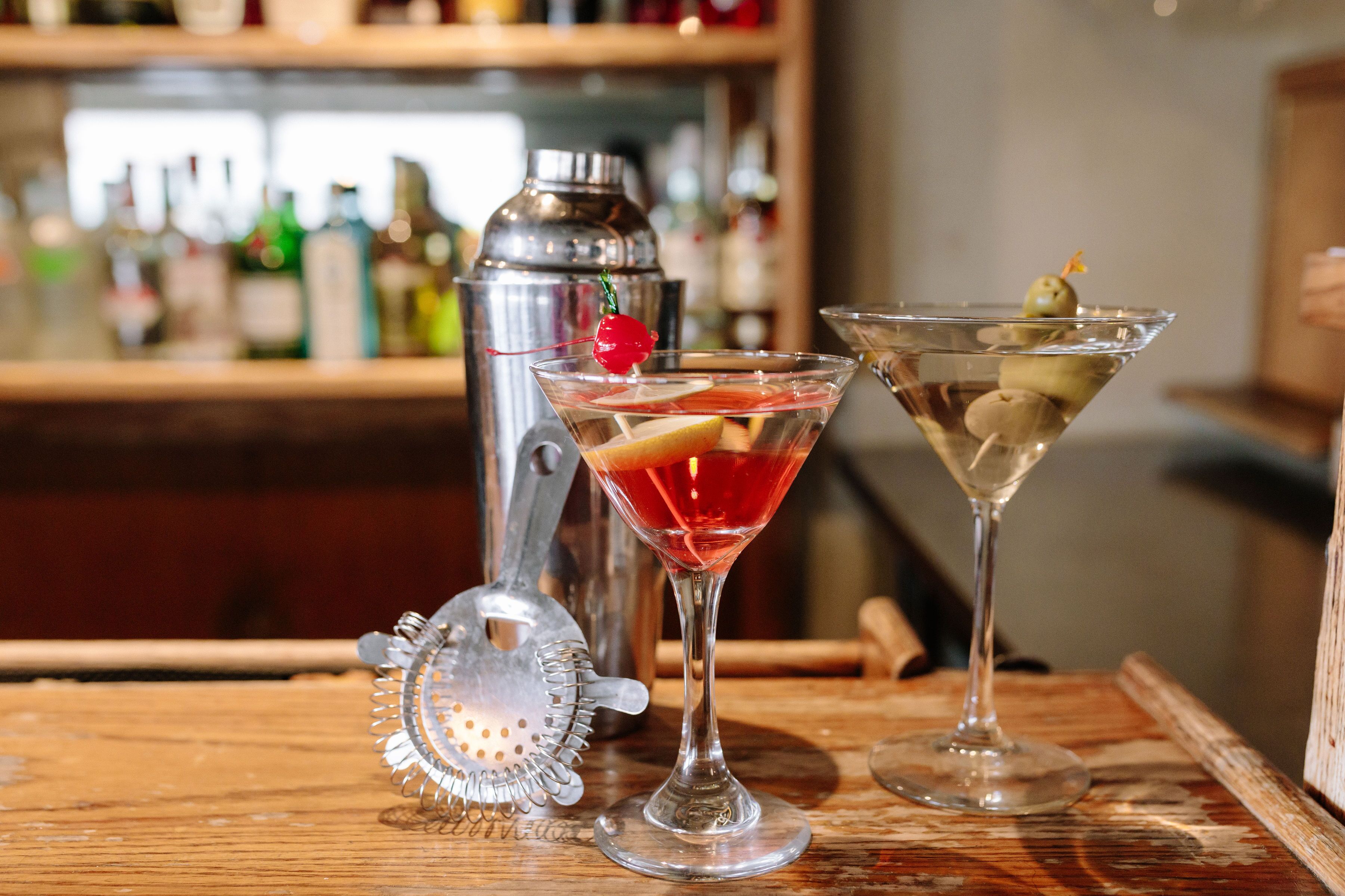 Deux verres à martini sur une surface en bois, avec un shaker à cocktails et une passoire en arrière-plan.