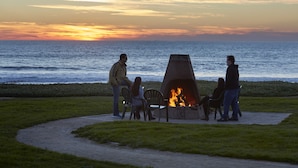 A group of people around a fire pit by the ocean at sunset.