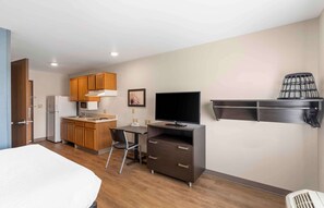 A compact hotel room with a kitchenette, a bed, a desk, a television, and a shelf with a basket.