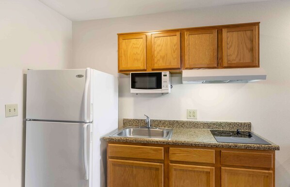 A compact kitchen with wooden cabinets, a stainless steel refrigerator, a microwave, and a sink with a granite countertop.