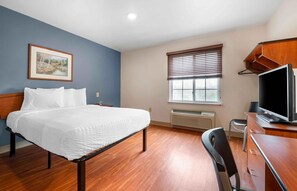 Habitación de hotel con cama, escritorio, silla, televisor, ventana con persianas, un cuadro en la pared y un radiador.