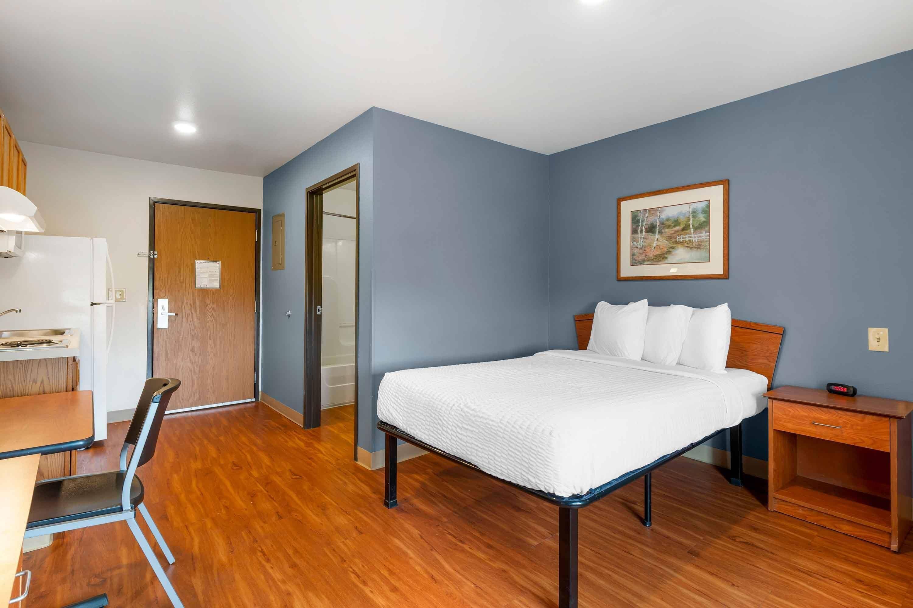 Quarto de hotel com uma cama, uma escrivaninha, uma cadeira, uma mesinha de cabeceira e um quadro na parede.