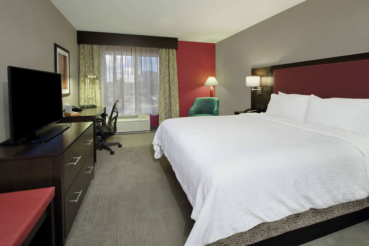 A hotel room with a bed, a desk with a TV, a chair, a lamp, and a window with curtains.
