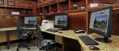 Zona de recepción de un hotel con tres estaciones de trabajo de computadora, cada una con monitor, teclado y ratón. Hay sillas de oficina, una impresora y un teléfono. El mostrador de recepción está flanqueado por armarios de madera con estantes superiores.