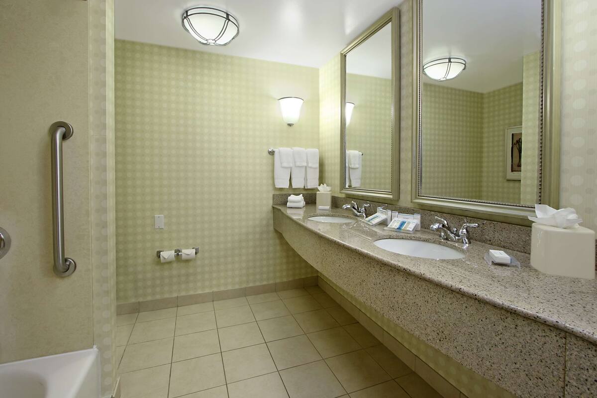 A hotel bathroom with a handrail, two sinks, a large mirror, and a towel rack.