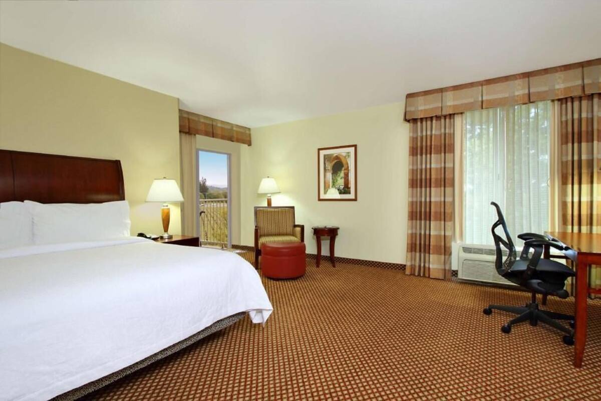 A hotel room with a large bed, two bedside tables with lamps, a chair, a desk with a chair, and a window with curtains.