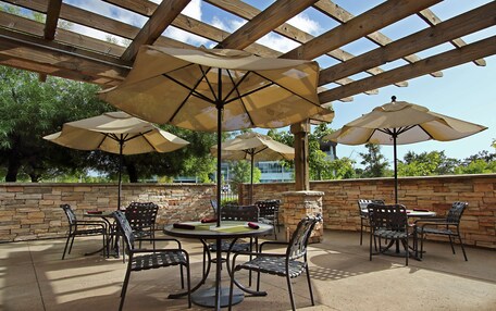 An outdoor patio with stone walls, wooden pergola, and multiple tables and chairs under beige umbrellas.