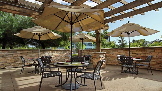 An outdoor patio with stone walls, wooden pergola, and multiple tables and chairs under beige umbrellas. - Hilton Garden Inn Sonoma County Airport (Santa Rosa)
