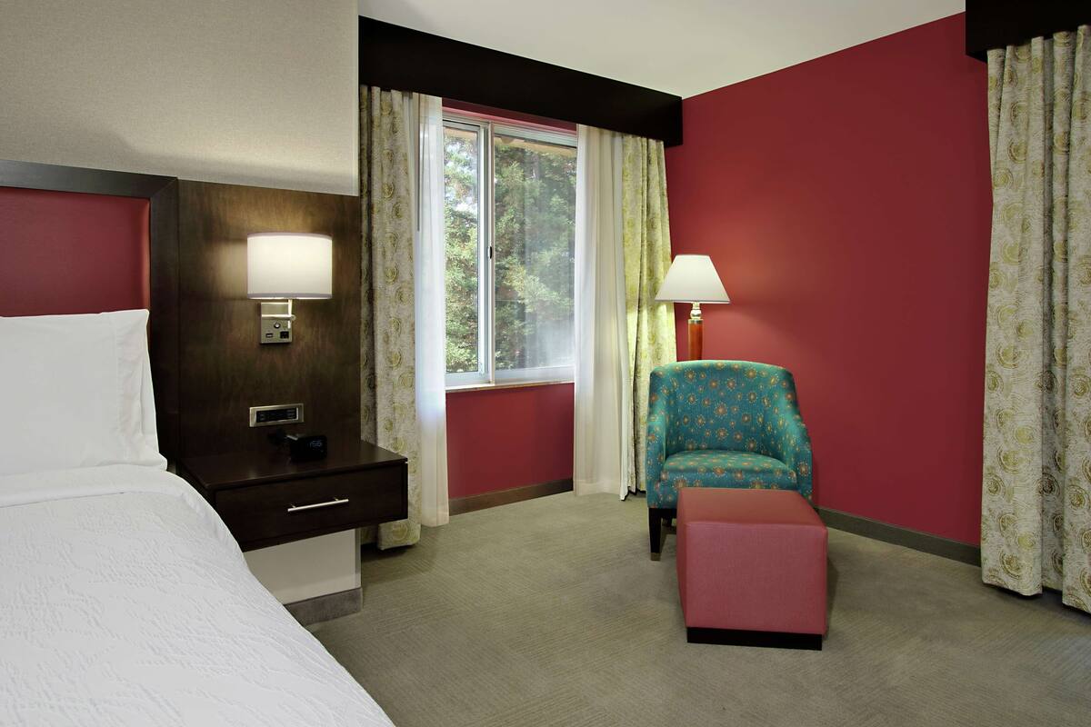 A hotel room with a bed, a desk with a lamp, a chair, and a window with curtains.