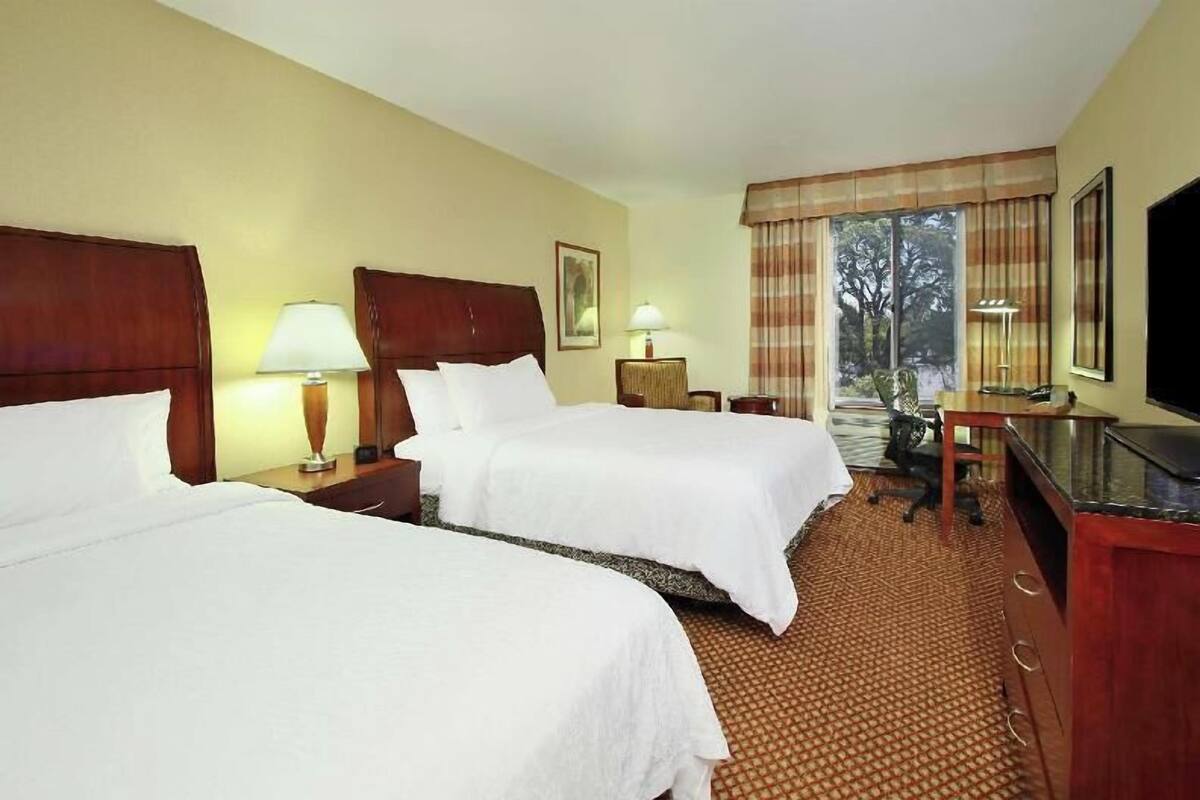 A hotel room with two beds, a desk, a chair, a TV, and a window with curtains.