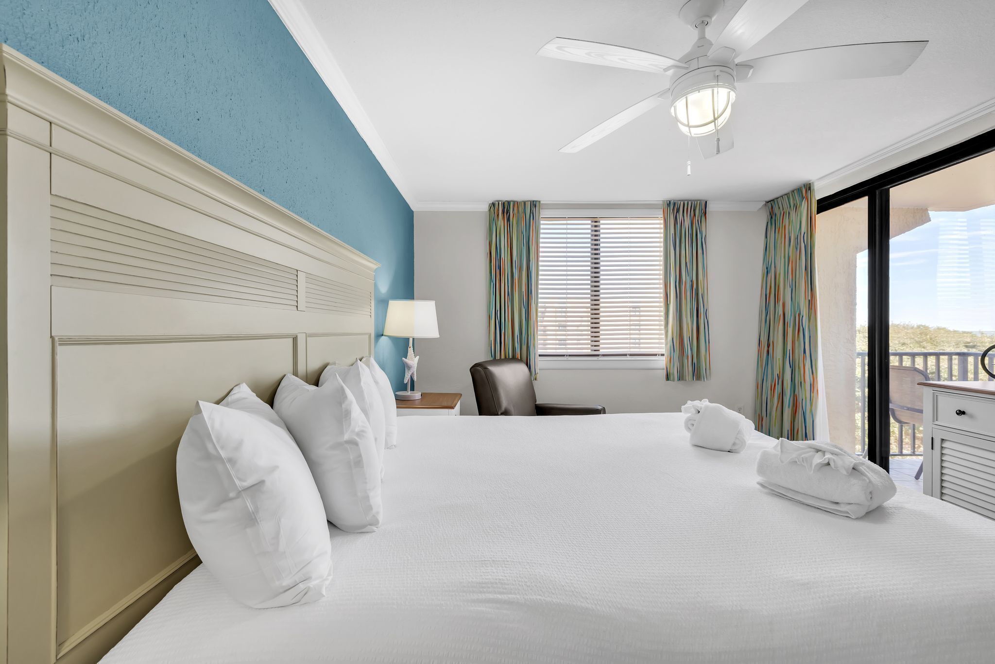 A hotel room with a large bed, a ceiling fan, a desk with a lamp, and a view of the outdoors through the window.