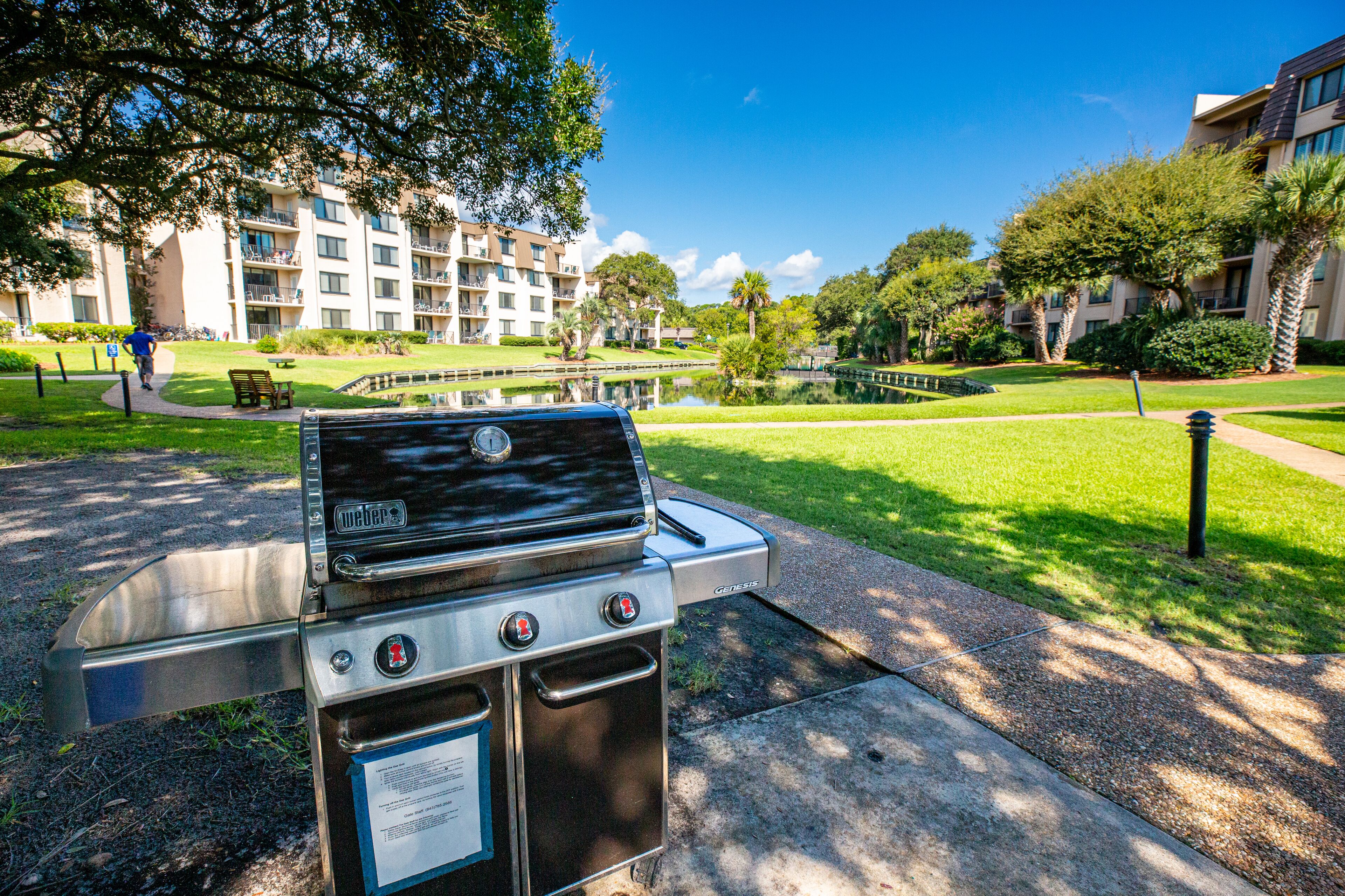 A Weber gas grill with a stainless steel finish, located in a well-maintained outdoor area with a pond, benches, and lush greenery.