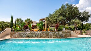 A swimming pool with a waterfall feature, surrounded by lush greenery and a building with a curved roof. - Seawatch at Island Club (Hilton Head Island)