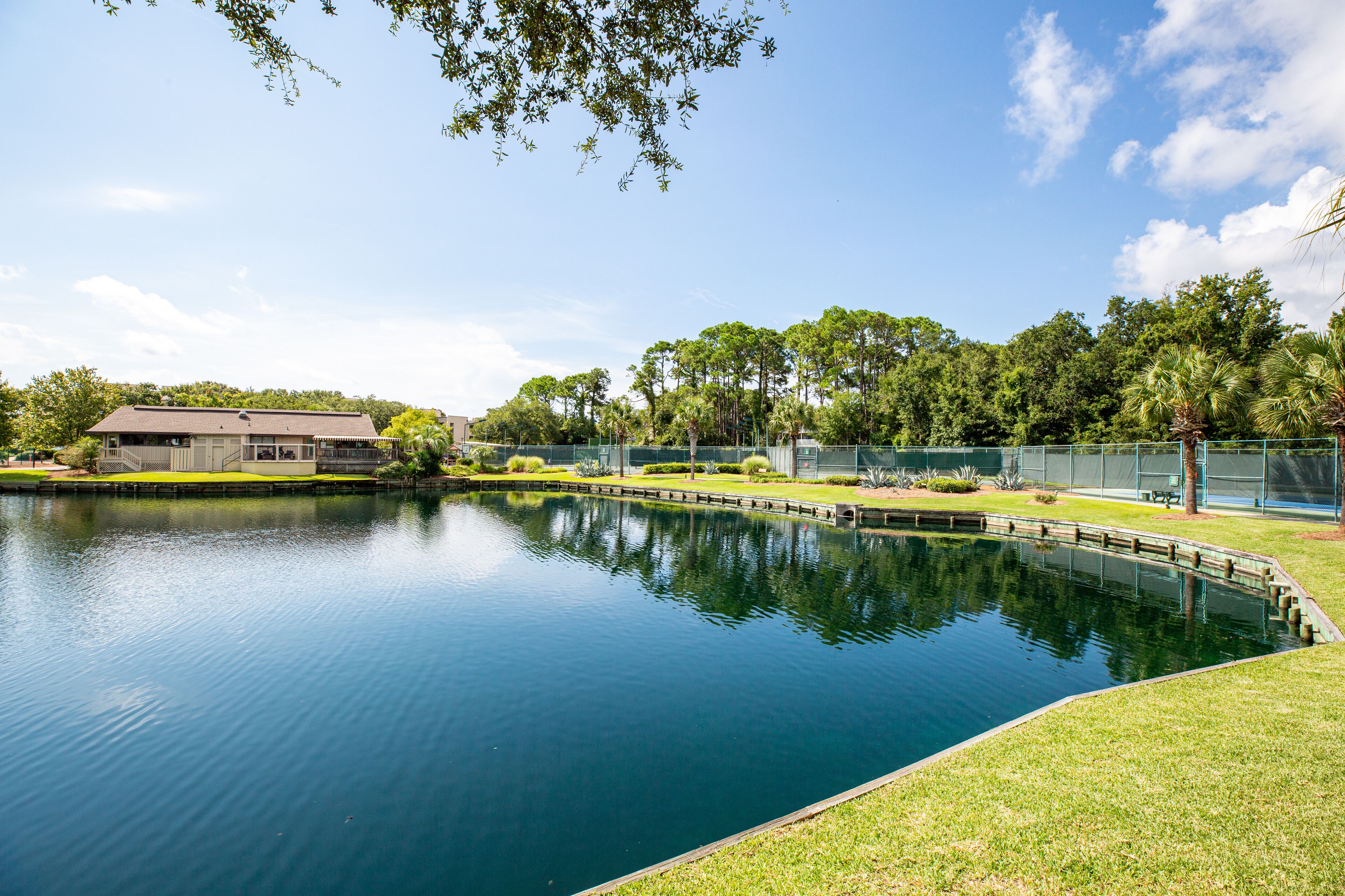 A calm lake with a grassy bank, surrounded by trees and a building with a flat roof.