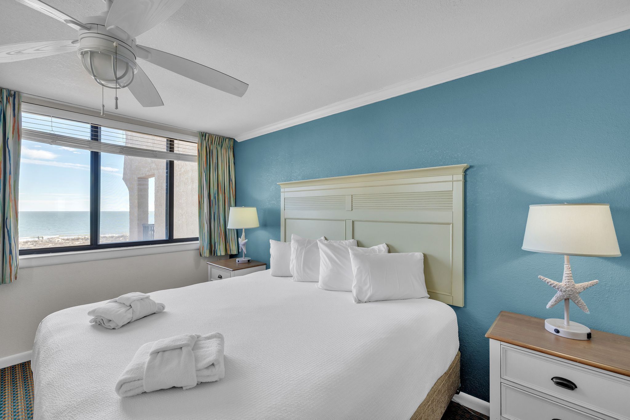 A hotel room with a bed, a nightstand, a lamp, a starfish decoration, and a view of the ocean through the window.