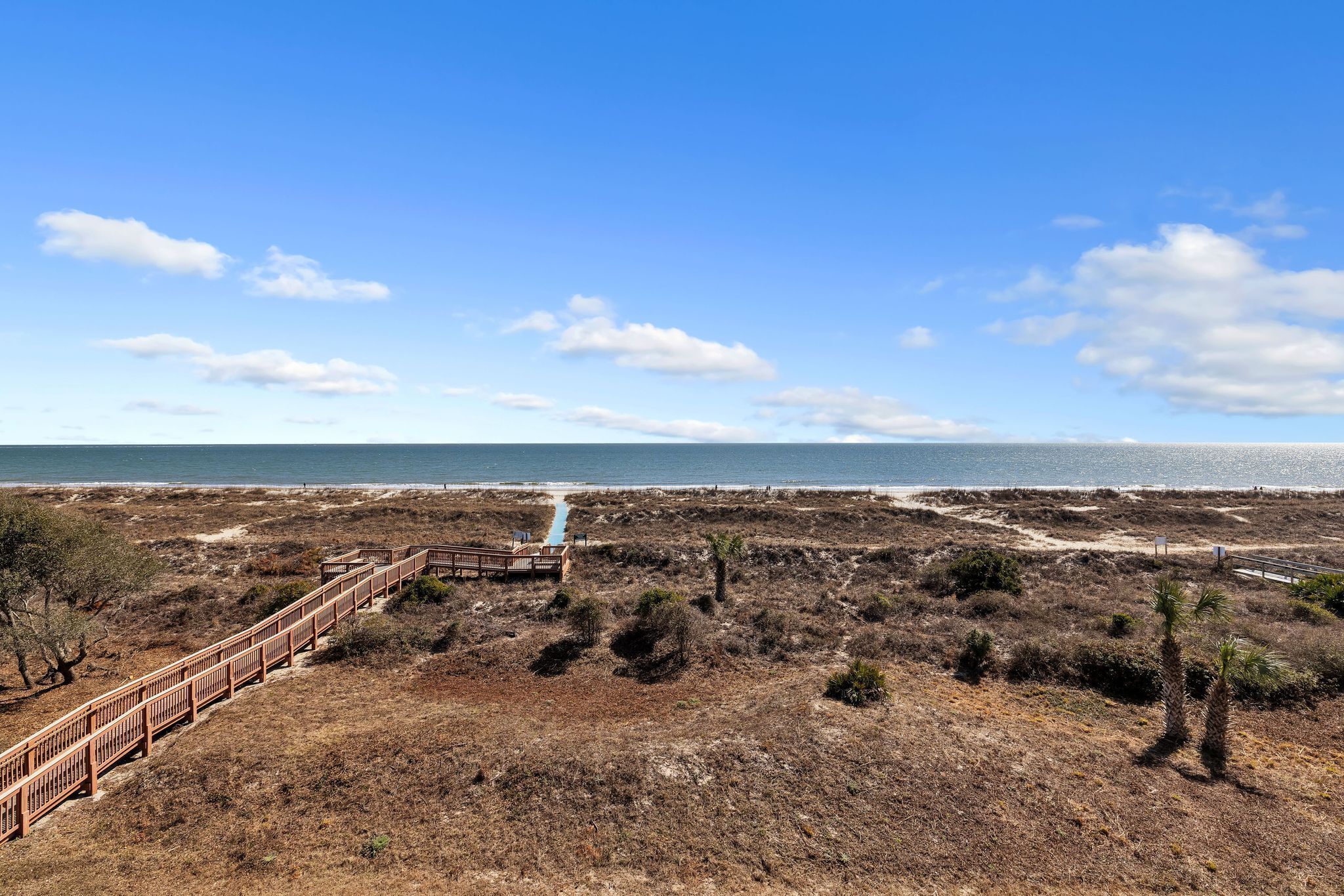 Un paisaje costero con un paseo marítimo de madera que conduce a una playa, rodeado de vegetación seca y un cielo despejado.