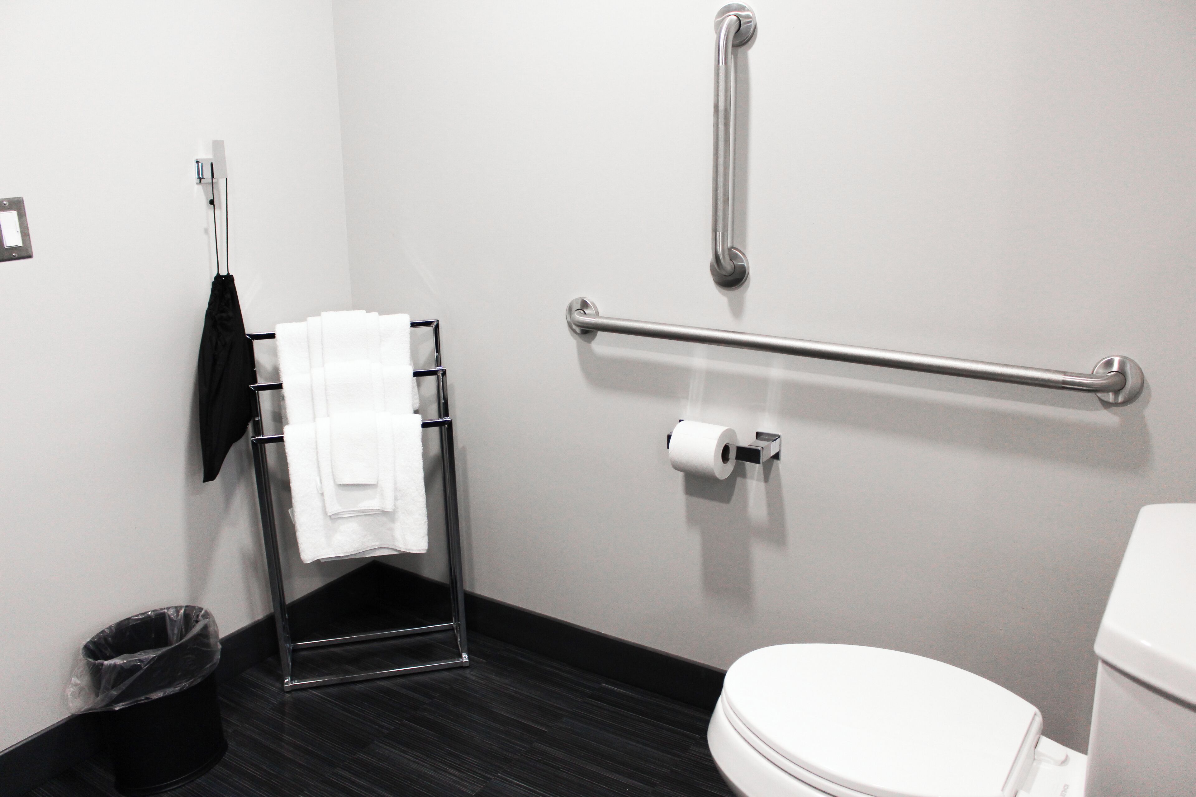 A modern bathroom with a white toilet, a towel rack with a white towel, a black trash bin, and a handrail.