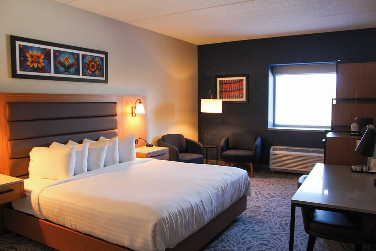 A hotel room with a large bed, two bedside tables, a desk, a chair, and a window with natural light.