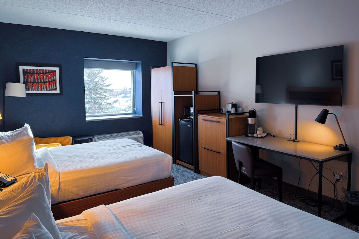 A hotel room with two beds, a desk, a TV, and a window with a view of trees.