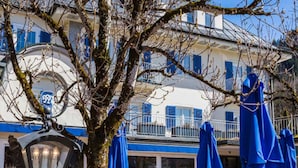 Een terras met tafels en stoelen, een grote boom, en een gebouw met blauwe luiken op de achtergrond.