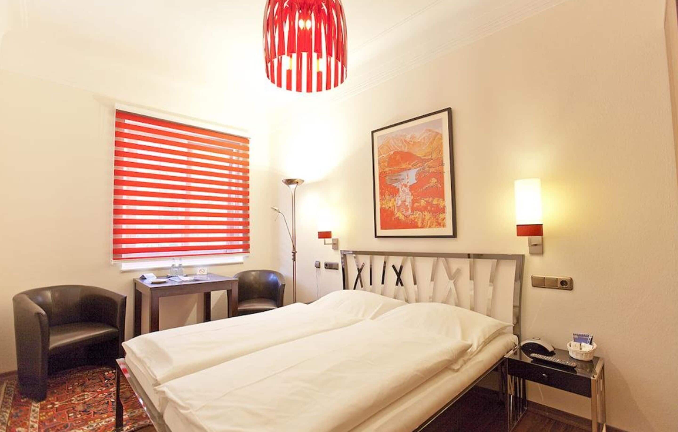 A hotel room with a bed, two bedside tables, a desk, a chair, a red pendant light, and a window with red blinds.