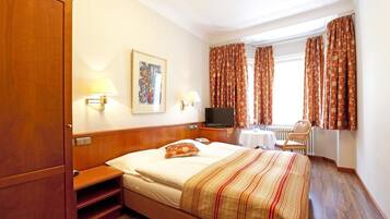 A hotel room with a bed, a TV, a desk with a chair, a window with curtains, and a painting on the wall.