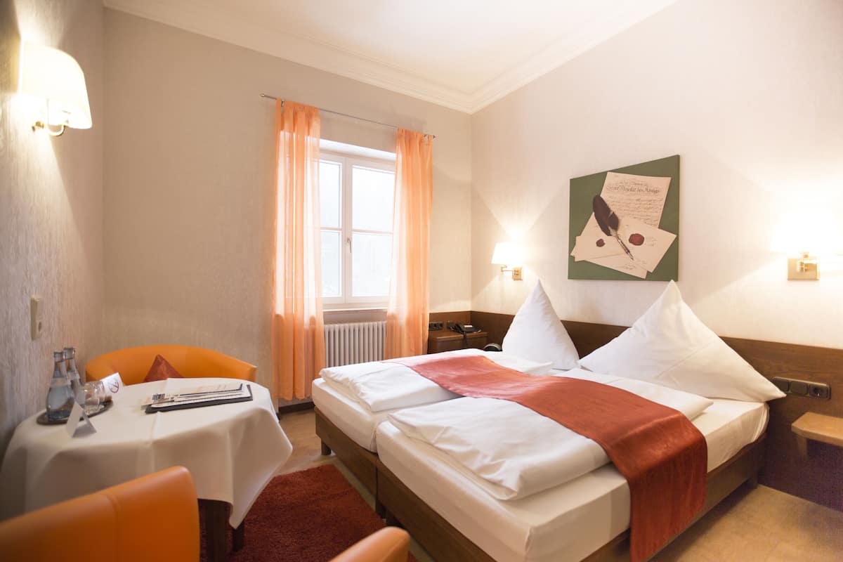 A hotel room with a bed, a table with a chair, a window with curtains, a wall-mounted lamp, and a framed picture on the wall.